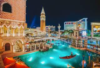 A couple in a gondola floating in the lit up canal at night in front of the Venetian hotel