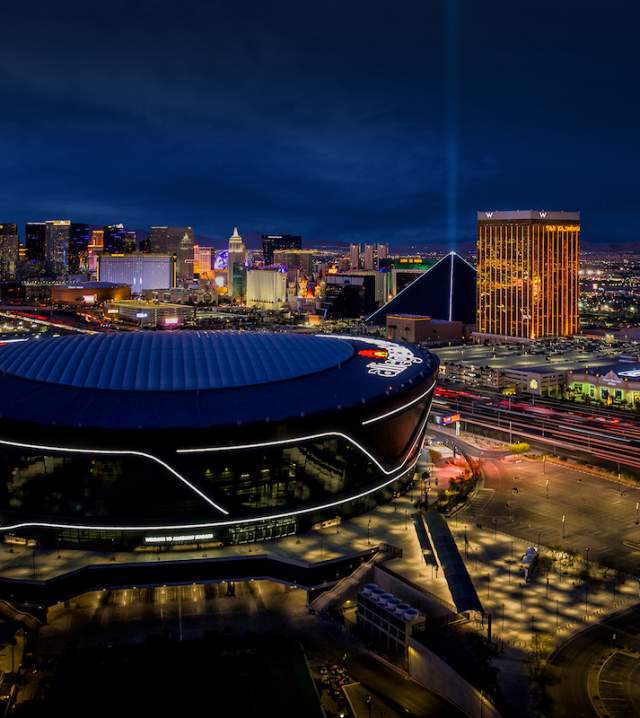 An aerial view of Allegiant Stadium at night with the Las Vegas Strip in the background.