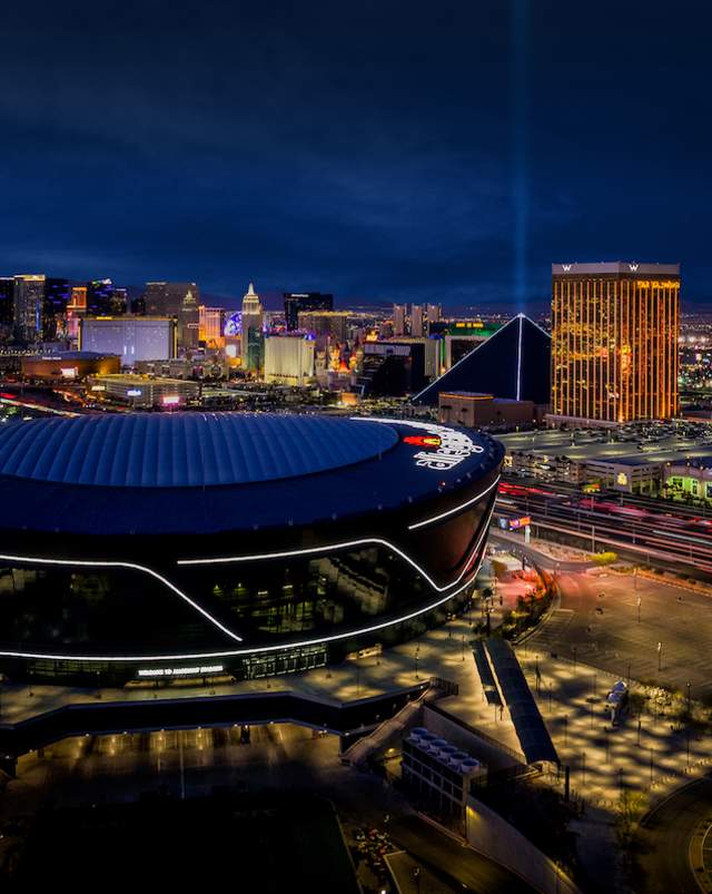 An aerial view of Allegiant Stadium at night with the Las Vegas Strip in the background.