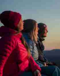 Tre mennesker sitter og nyter utsikten fra Gaustatoppen i Telemark i skumringen