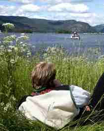 Ein in der Nationaltracht Setesdalbunad liegt im Gras und und schaut auf ein Boot am Byglandsfjord, Setesdal
