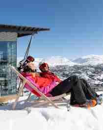 Two people in skiing clothes sit in deck chairs outside Skarsnuten Hotel and enjoy the sunny weather and the wide views of the mountains of Hemsedal, Eastern Norway
