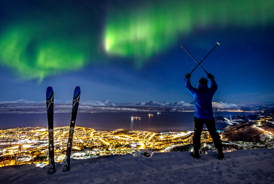 A man alpine skiing over Narvik town while spectacular northern lights