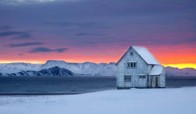 A wooden house next to the water on a snow-covered landscape with sunset colors in the background at Hasvik in Finnmark, Northern Norway