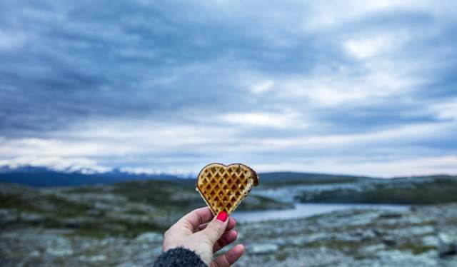 A hand holding up a heart-shaped waffle with beautiful scenery in the background