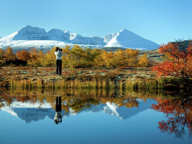 A woman taking photos of a lake and trees in autumn colours in the Rondane national park, Eastern Norway. Snowclad mountains in the background.