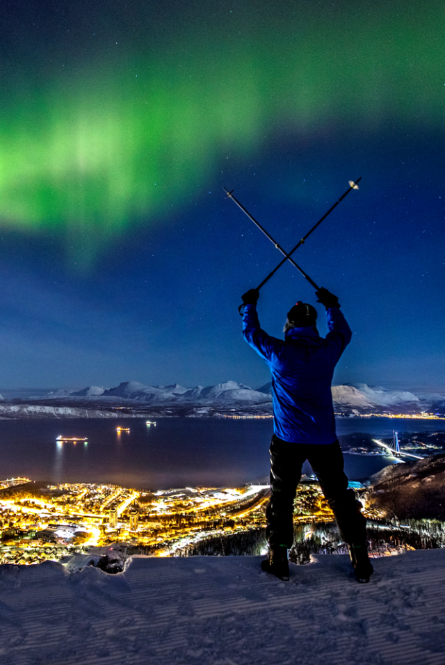 A man alpine skiing over Narvik town while spectacular northern lights