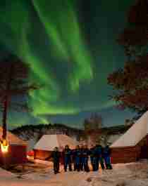 A group of people standing in front of several cabins under the northern lights