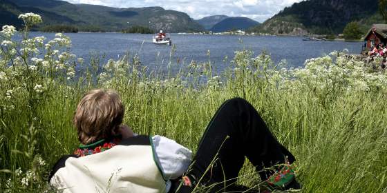 A man in the national costume Setesdalsbunad lying in the grass and watching an old steamboat on the Byglandsfjord in Setesdal, Southern Norway