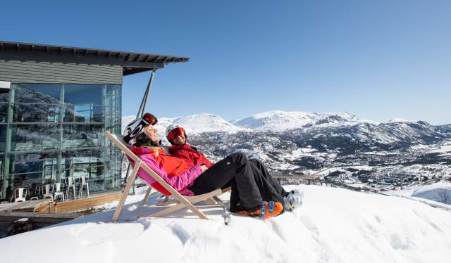 Two people in skiing clothes sit in deck chairs outside Skarsnuten Hotel and enjoy the sunny weather and the wide views of the mountains of Hemsedal, Eastern Norway