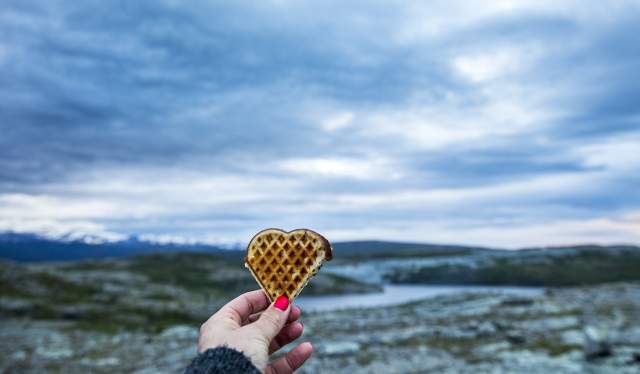 A hand holding op a heart-shaped waffle with beautiful scenery in the background