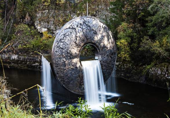A round metal sculpture surrounded by nature, with water running through at Kistefos in Jevnaker