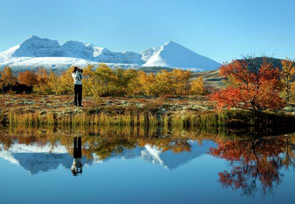 A woman taking photos of a lake and trees in autumn colours in the Rondane national park, Eastern Norway. Snowclad mountains in the background.