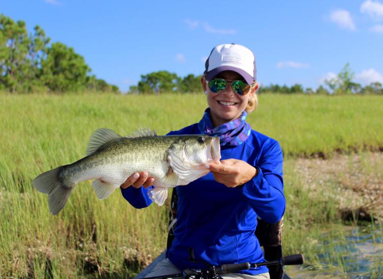 a woman in a blue fishing shirt holds open the mouth of a bass she has caught