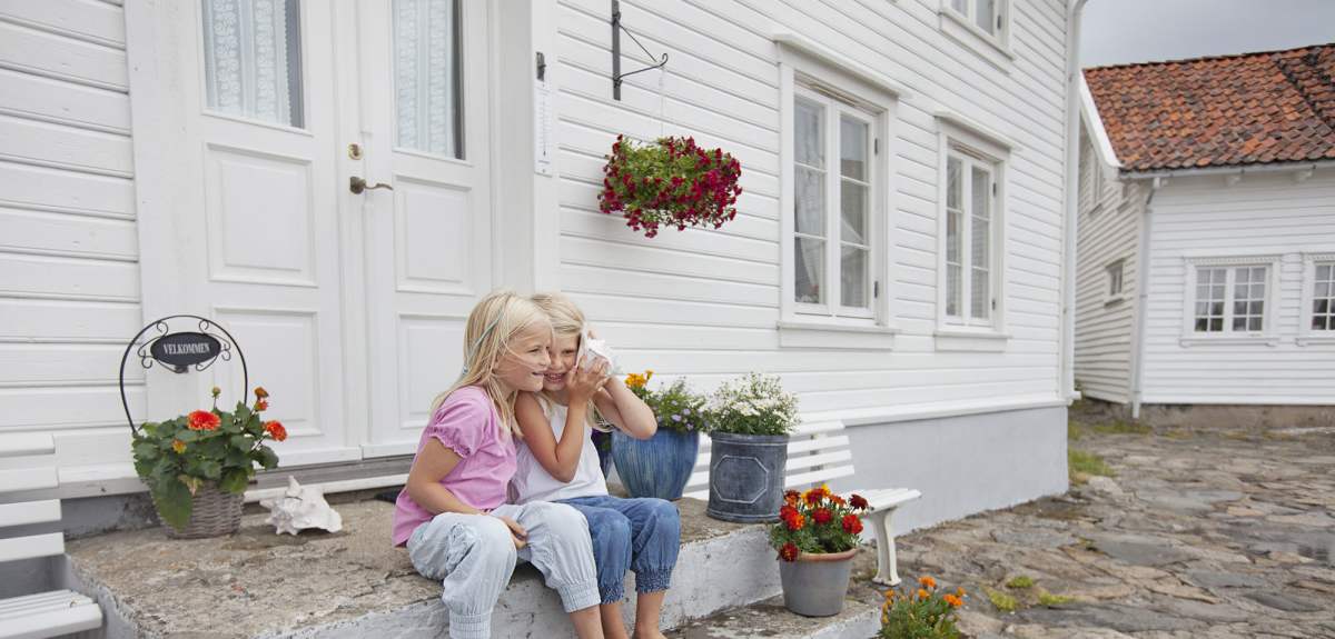 Two young girls talking while sitting on the stone steps in front of a white wooden house in Loshavn, Farsund, Southern Norway.