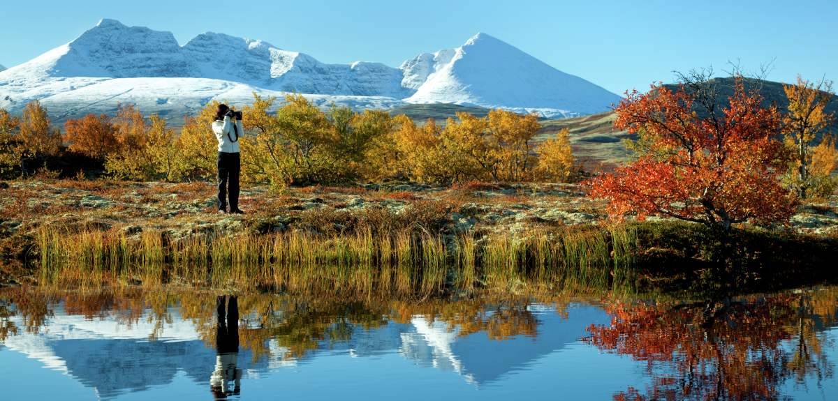 Una mujer hace fotos frente a un lago y varios árboles de colores otoñales en el Parque nacional de Rondane, Este de Noruega. Al fondo, montañas cubiertas de nieve.