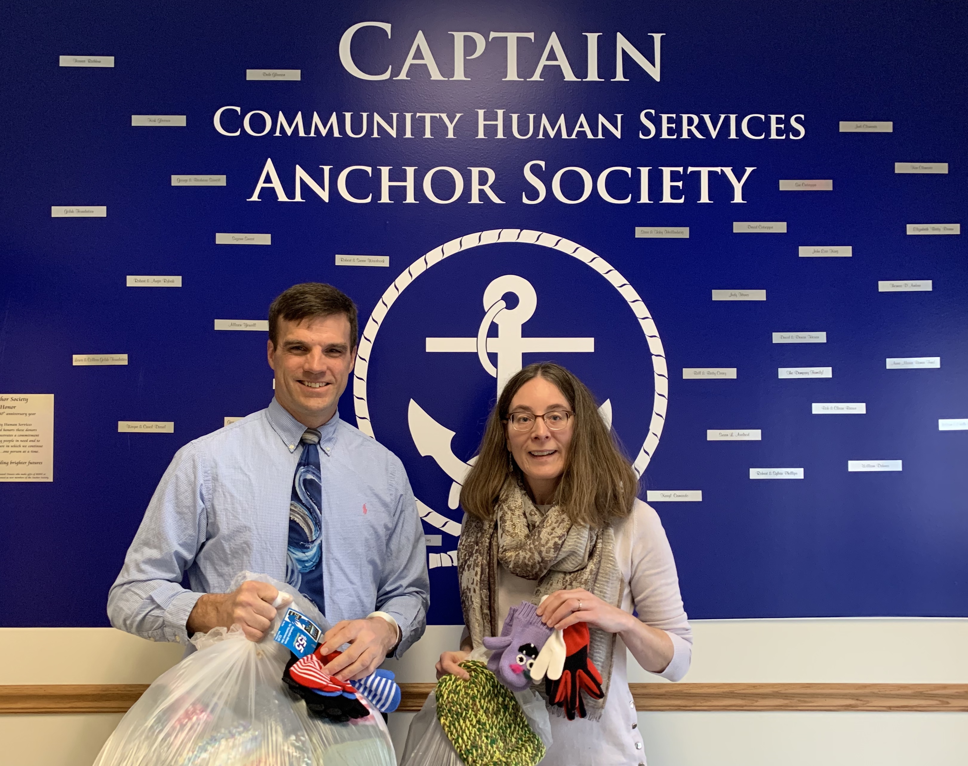 Woman and man holding bag of mittens and hats in front of Captain Community Human Resources Anchor Society sign